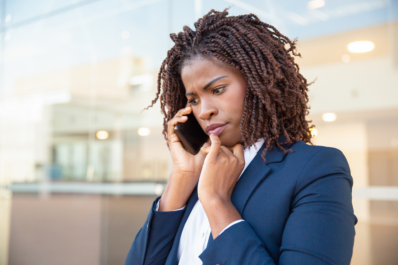 Frowning concerned manager speaking on cellphone Frowning concerned manager speaking on cellphone outside. Young African American business woman standing near outdoor glass wall. Mobile phone talk concept
